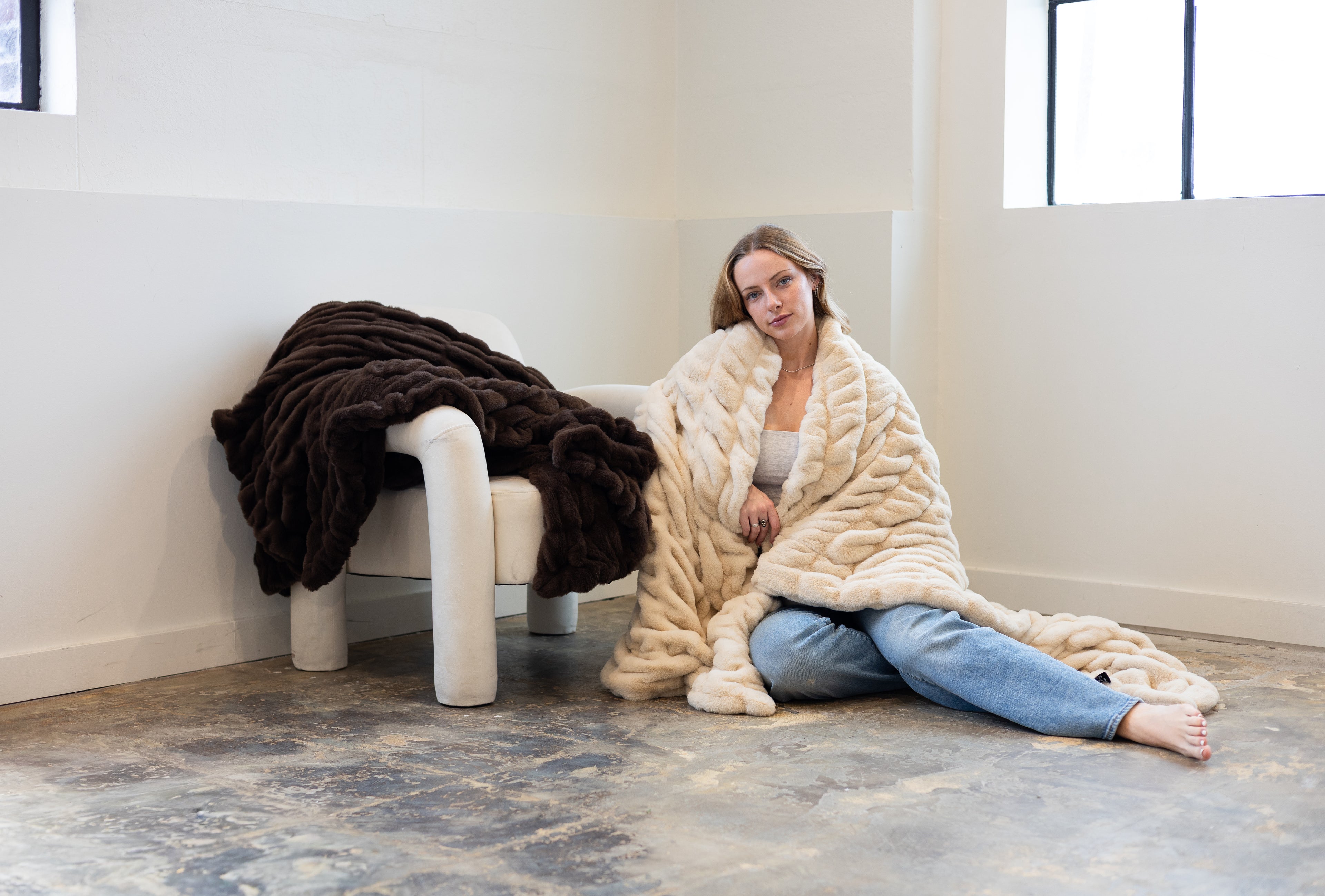Woman sitting on the floor wrapped in a large beige blanket next to a chair with a dark blanket.